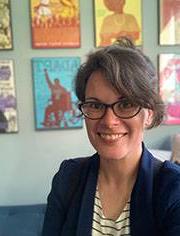 Allison Brewer, headshot. They are wearing a navy jacket over a blue and white striped top and glasses and smiling at the camera.