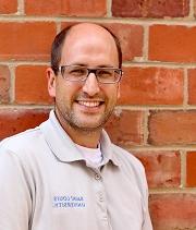 Bobby Wassel, headshot, wearing a grey 博彩网址大全 polo shirt, stands in front of a brick wall and smiles.