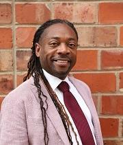 Richard Marks, wearing a pink suit, maroon tie and white shirt, stands in front of a brick wall and smiles